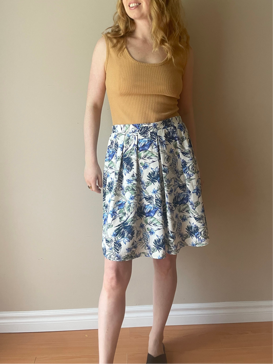 Woman wearing Skies Are Blue pleated skirt with white base and blue tropical floral print, styled with a camel ribbed sleeveless tank for a summer-ready look.