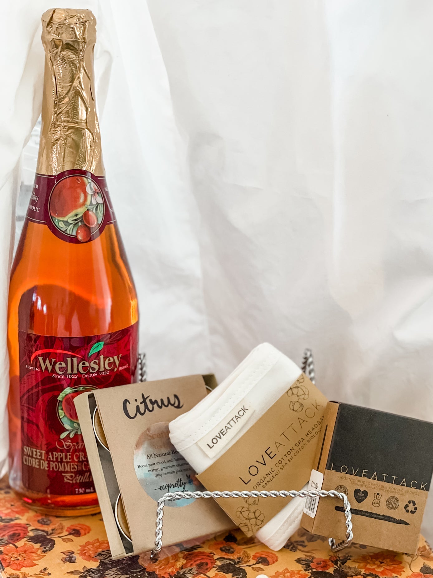 A gift bundle experience set including a bottle of Wellesley non-alcoholic sparkling cider, a pack of scented beeswax tealight candles, a spa headband, a Konjac sponge, all presented in an eco-friendly box.