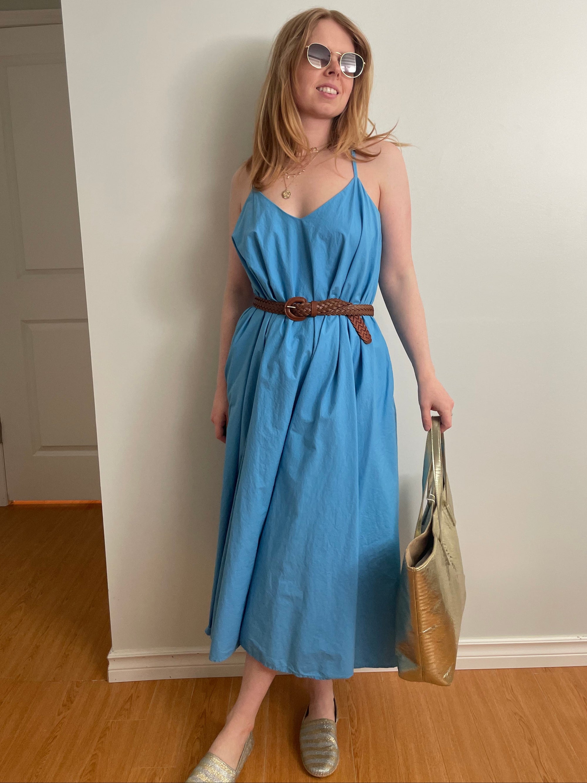 A woman wearing a light blue midi dress with thin straps and a brown woven belt, standing in a hallway and holding a large gold handbag.