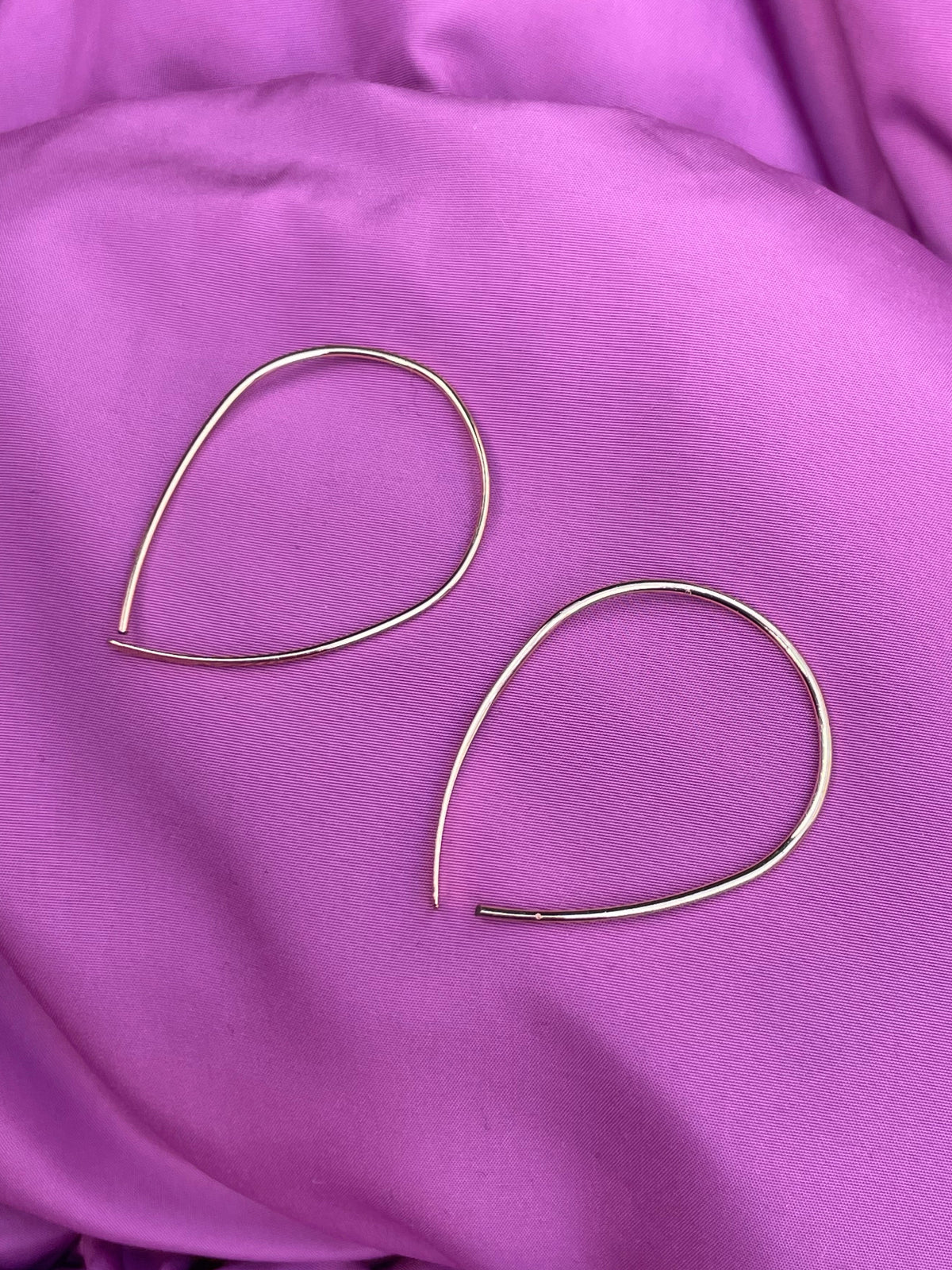 Two thin gold-toned teardrop hoop earrings resting on draped purple fabric.