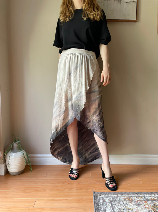 Model wearing a blush and grey dip-dye high-low silk skirt by Gypsy 05 with a black rolled sleeve tee and black heeled sandals in a softly lit room.