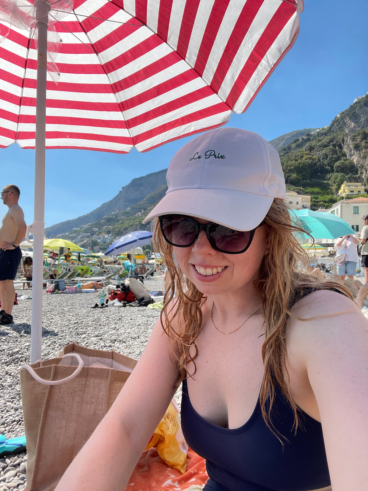A woman on a beach in Amalfi Italy wearing a white baseball cap with 'Le Prix' embroidered on it, dark sunglasses, and a navy swimsuit, under a red and white striped beach umbrella with mountains in the background.