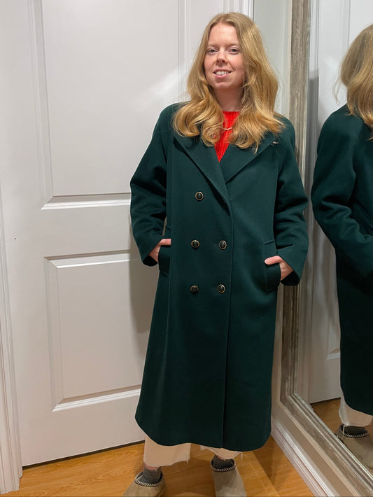 Woman wearing a dark green coat standing in front of a mirror.