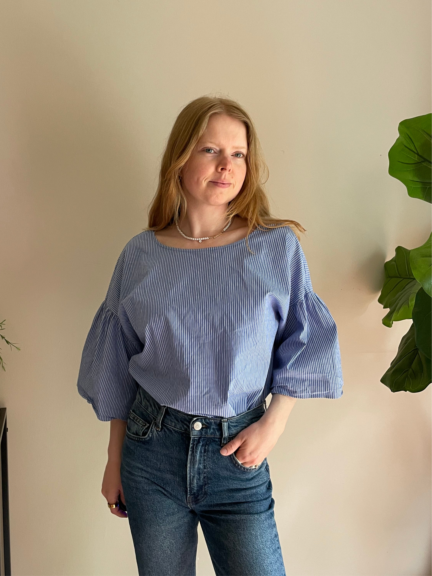 Woman wearing a blue striped blouse and jeans against a beige wall with a plant.