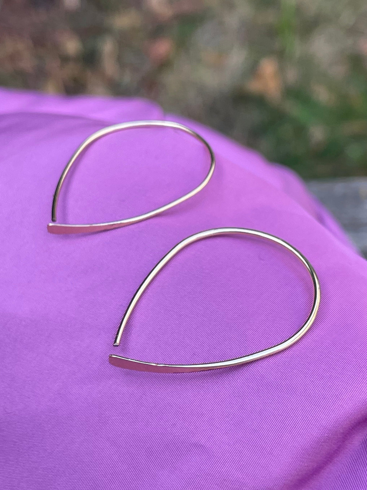 A pair of thin, open teardrop-shaped hoop earrings in a polished gold-tone metal, resting on a draped purple fabric surface with an outdoor blurred background.