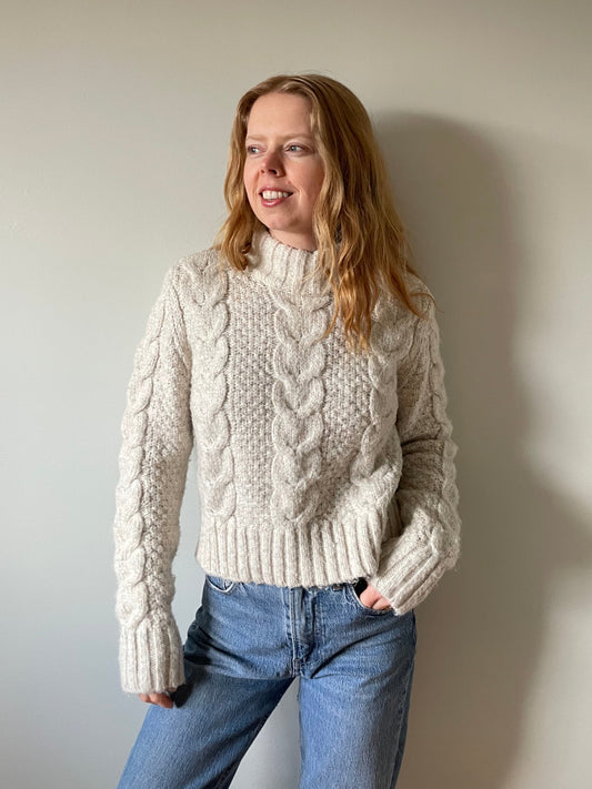 Model wearing a cream cable knit mock neck sweater with long sleeves and ribbed trim, styled with blue jeans against a neutral background.