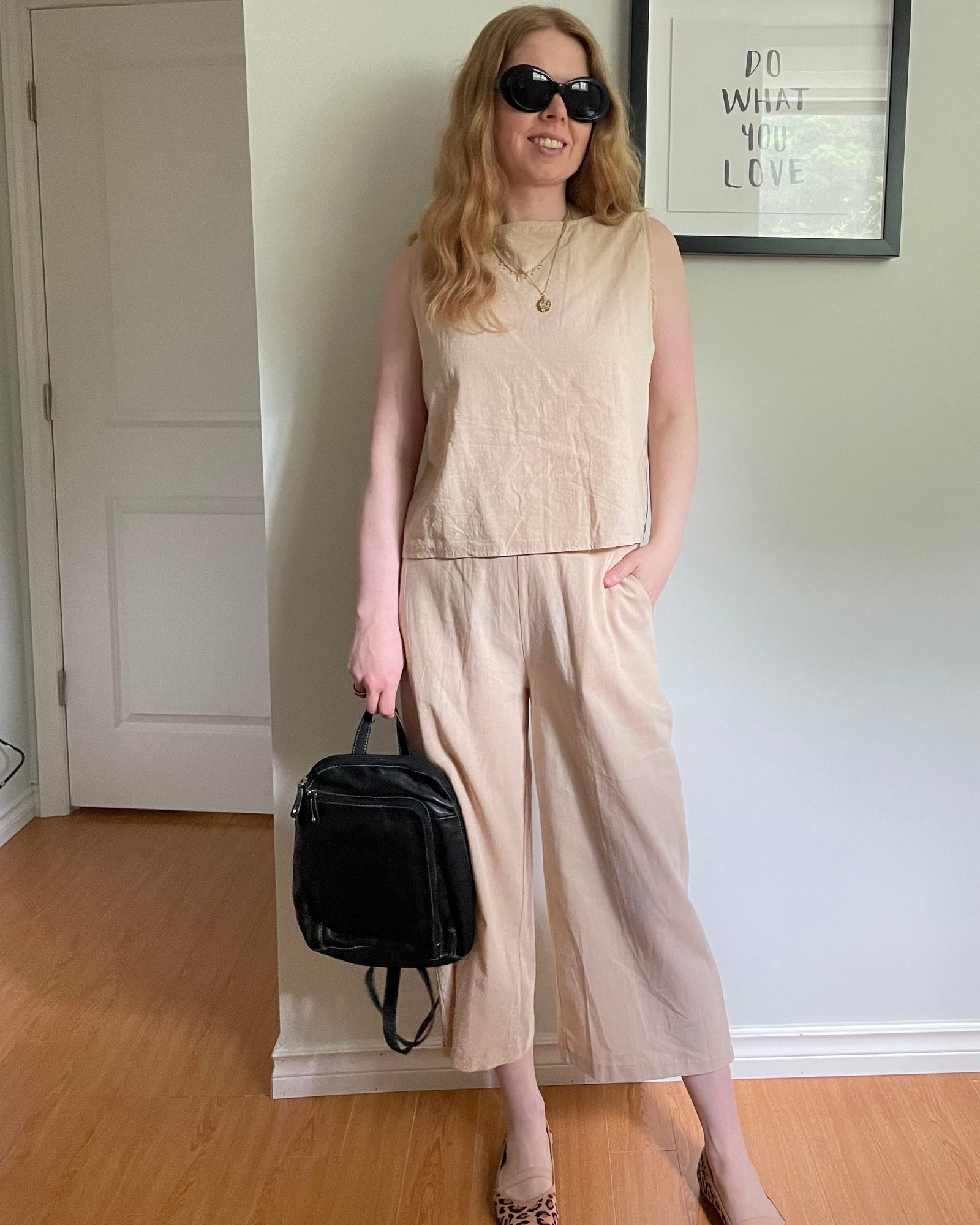 A woman wearing a beige sleeveless wide-leg jumpsuit and leopard print heels, holding a black backpack, standing in an indoor room with a light-colored wall and wood floor.