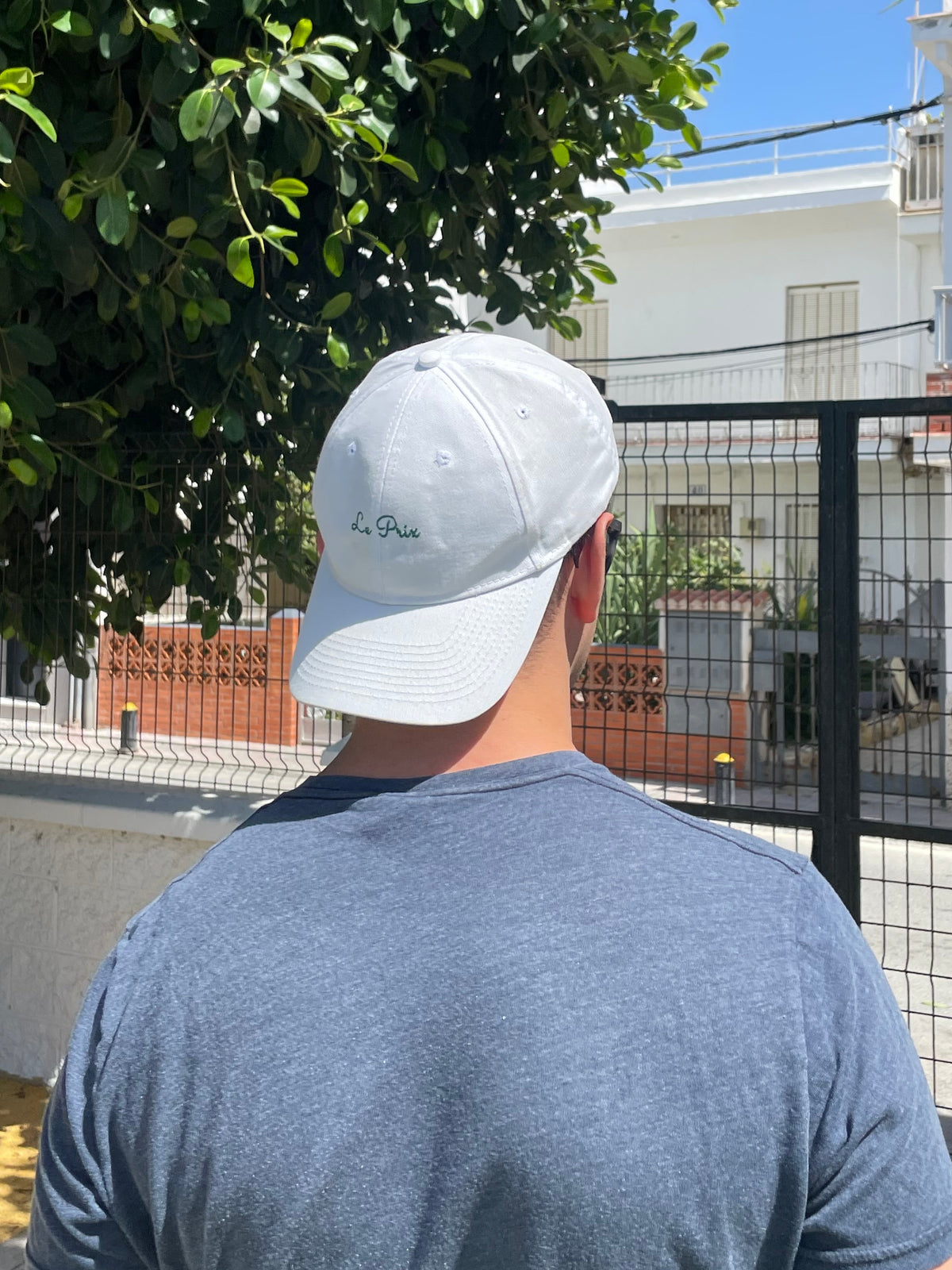 A man viewed from behind wearing a white baseball cap with a small black script logo and a heathered blue-gray t-shirt, standing outdoors in Spain in front of a white building and green trees.